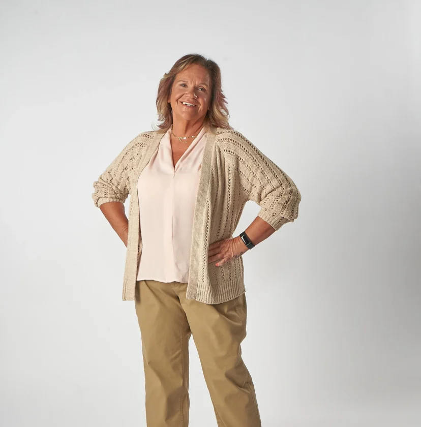 A woman stands smiling with her hands on her hips, wearing a beige cardigan over a light pink blouse and khaki pants, in front of a plain white background.