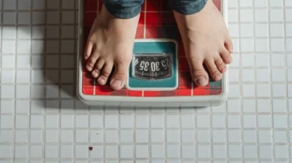 Person standing on a red analog bathroom scale, displaying a weight measurement, on a white tiled floor.