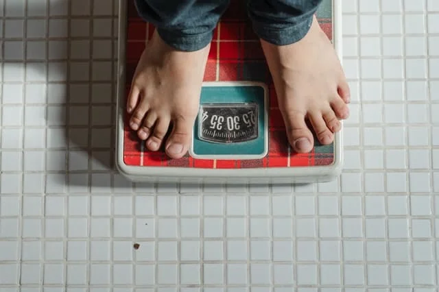 Person standing on a red analog bathroom scale, displaying a weight measurement, on a white tiled floor.