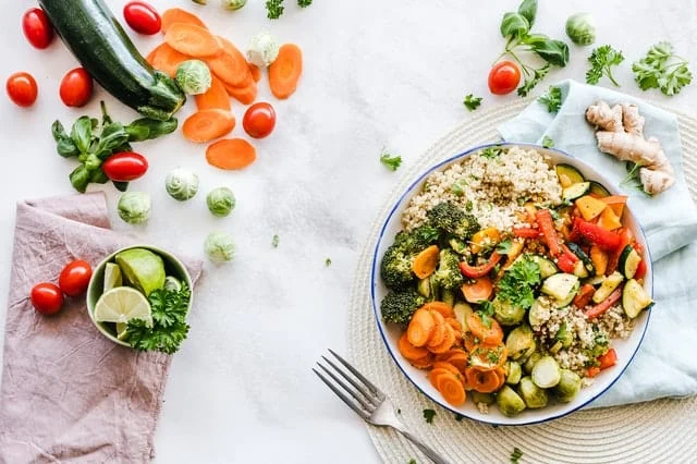 A bowl of quinoa with broccoli, carrots, zucchini, and red peppers is served on a placemat, surrounded by fresh vegetables and lime wedges on a white surface.