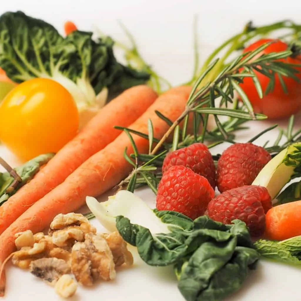 Assorted fresh vegetables, raspberries, walnuts, and rosemary arranged on a white surface.
