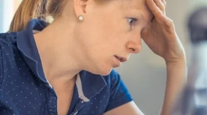 A woman with a ponytail sits at a desk, looking at a laptop with a concerned expression and her hand on her forehead. A glass of water and a bottle are in the foreground.
