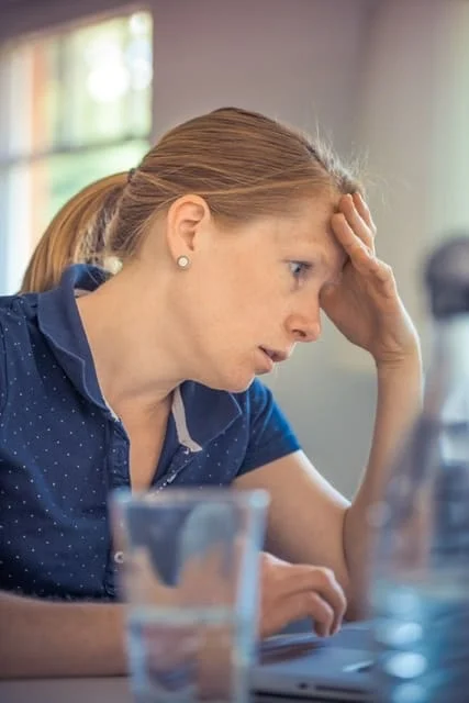 A woman with a ponytail sits at a desk, looking at a laptop with a concerned expression and her hand on her forehead. A glass of water and a bottle are in the foreground.