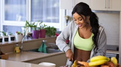 A woman in a green sports bra and gray hoodie smiles while cutting fruit in a bright kitchen, with bananas, sweet potatoes, and a bowl of berries on the counter.