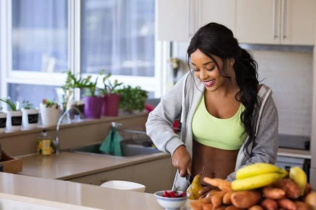 A woman in a green sports bra and gray hoodie smiles while cutting fruit in a bright kitchen, with bananas, sweet potatoes, and a bowl of berries on the counter.