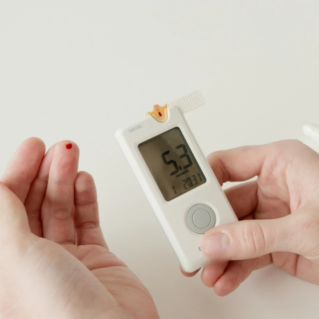 A person holds a blood glucose meter showing a reading of 5.3 mmol/L while a drop of blood is visible on their finger.