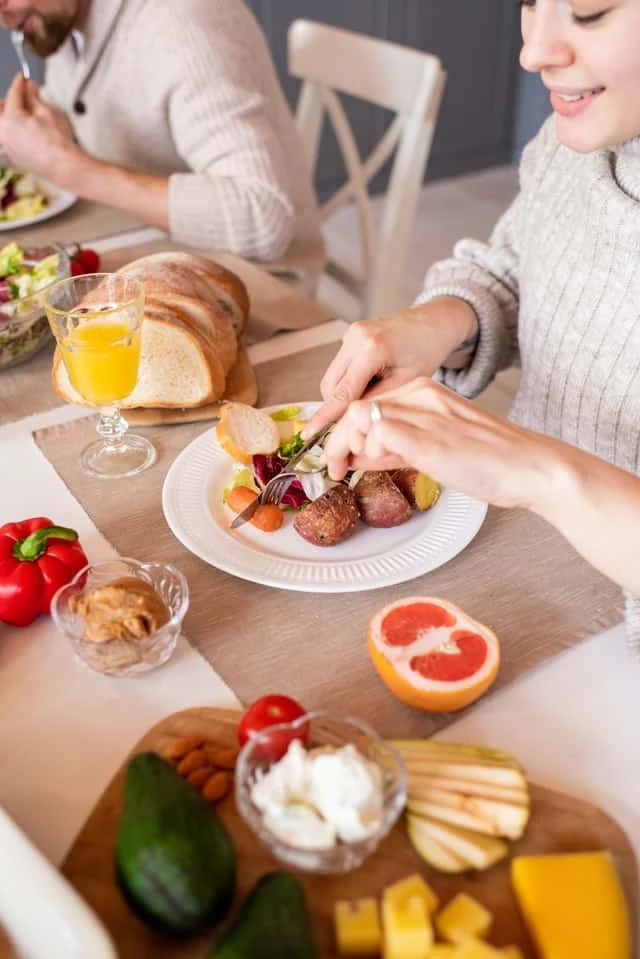 Two people sit at a table eating a meal with salad, bread, meat, grapefruit, avocado, cheese, and orange juice. The focus is on the food and the woman’s hands using a fork and knife.