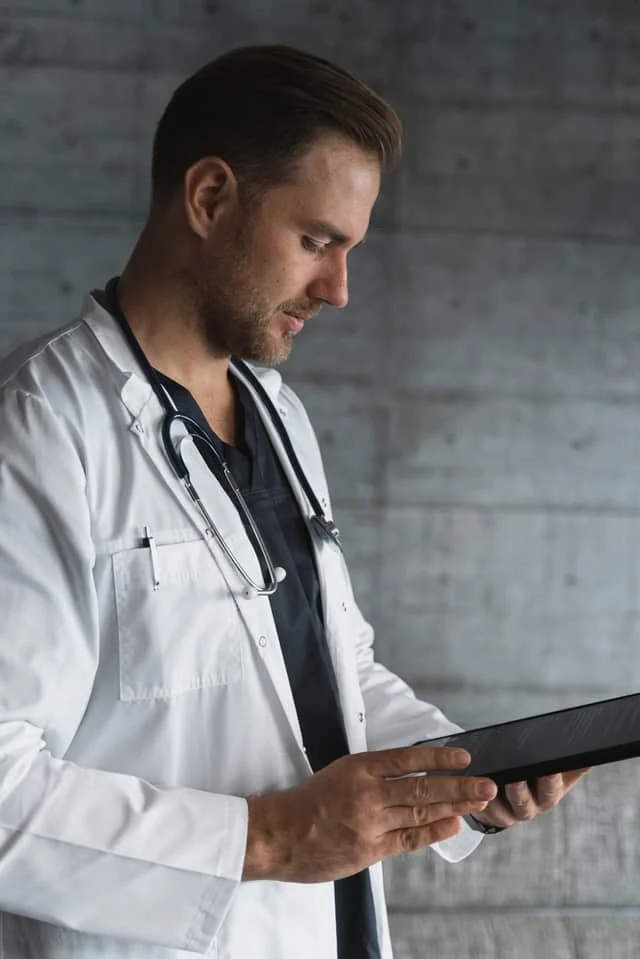 A male doctor wearing a white coat and stethoscope looks at a tablet device, standing against a gray textured wall.