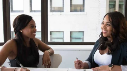 Two women sit at a table in an office, smiling and holding pens, with notebooks open in front of them.