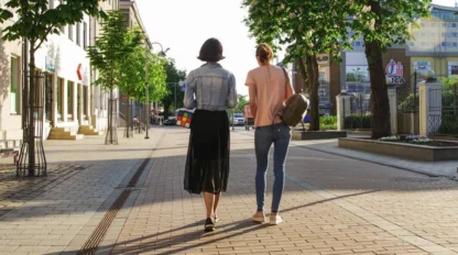 Two women walk down a sunlit, tree-lined city street, viewed from behind, one carrying a Rubik's cube bag and the other with a backpack.