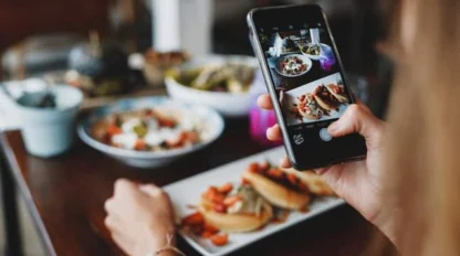 A person takes a photo of plated food with a smartphone at a table set with various dishes.