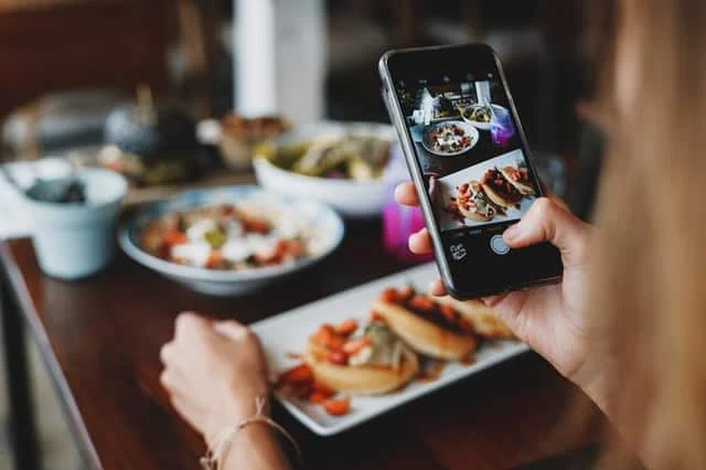 A person takes a photo of plated food with a smartphone at a table set with various dishes.