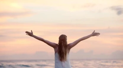 A woman with long hair stands facing the ocean with her arms outstretched, wearing a white dress, as the sun sets, casting a warm glow over the sky and water.
