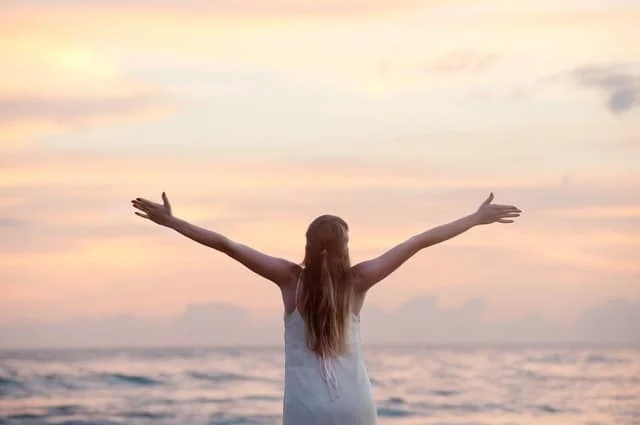 A woman with long hair stands facing the ocean with her arms outstretched, wearing a white dress, as the sun sets, casting a warm glow over the sky and water.