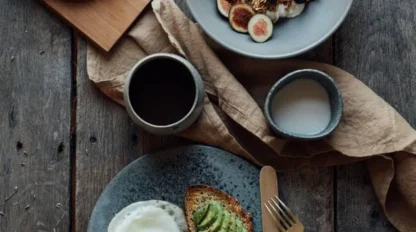 A breakfast spread with seeded bread slices, a bowl of granola with figs and blueberries, a cup of black coffee, a small bowl of milk, and avocado toast with a fried egg.