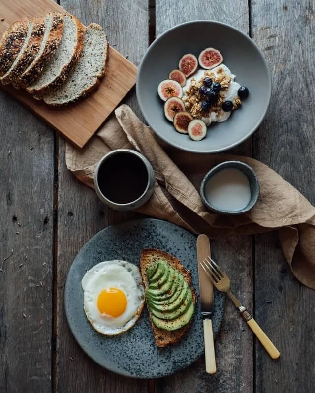A breakfast spread with seeded bread slices, a bowl of granola with figs and blueberries, a cup of black coffee, a small bowl of milk, and avocado toast with a fried egg.