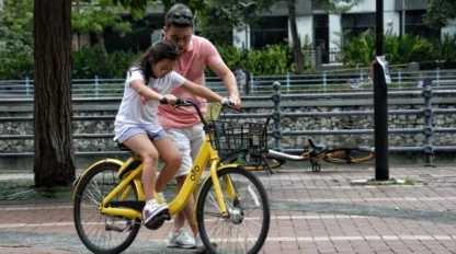 An adult helps a child learn to ride a yellow bicycle in an outdoor urban area with trees and a parked bike nearby.