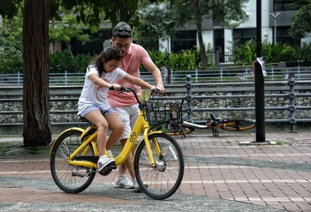 An adult helps a child learn to ride a yellow bicycle in an outdoor urban area with trees and a parked bike nearby.