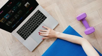 Person using a laptop on a wooden floor next to a blue exercise mat and a purple dumbbell.