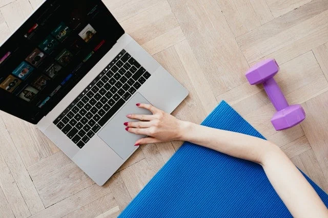 Person using a laptop on a wooden floor next to a blue exercise mat and a purple dumbbell.