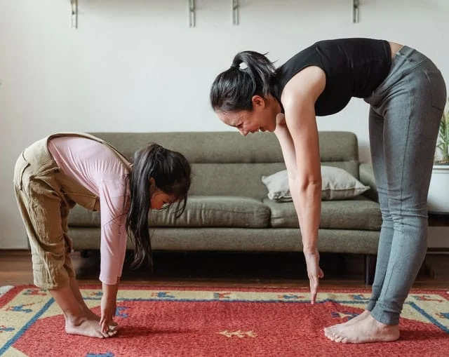 An adult and a child stand on a red carpet in a living room, bending forward with arms extended towards the floor, engaging in a stretching exercise together.
