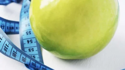 A green apple sits on a white surface with a blue measuring tape coiled around it.