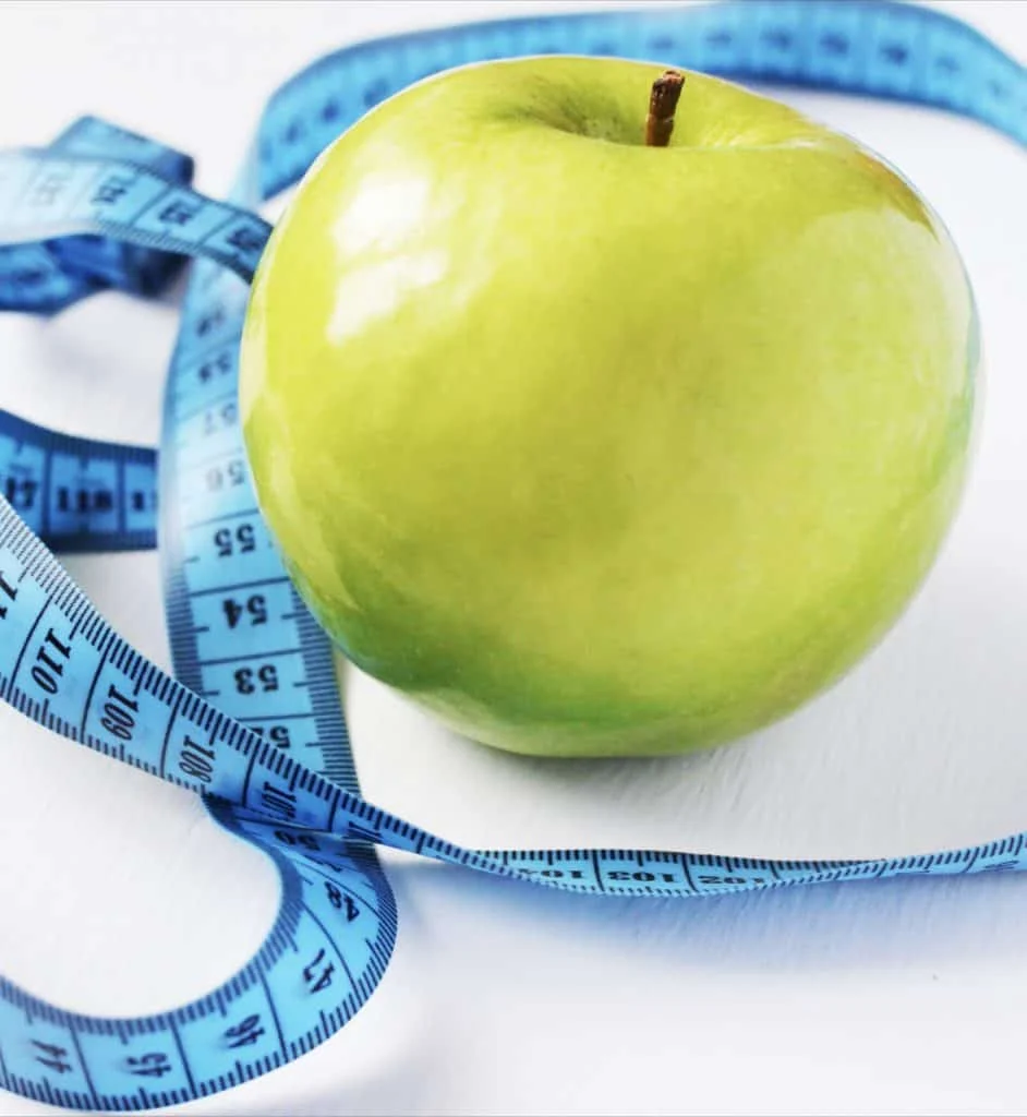 A green apple sits on a white surface with a blue measuring tape coiled around it.