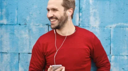 A man wearing a red sweater stands in front of a blue brick wall, holding a smartphone and smiling while listening to earphones.