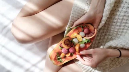 A person in a cozy sweater sits on a bed with crossed legs, holding a colorful fruit-topped toast. Sunlight filters through blinds, casting soft shadows across the scene.