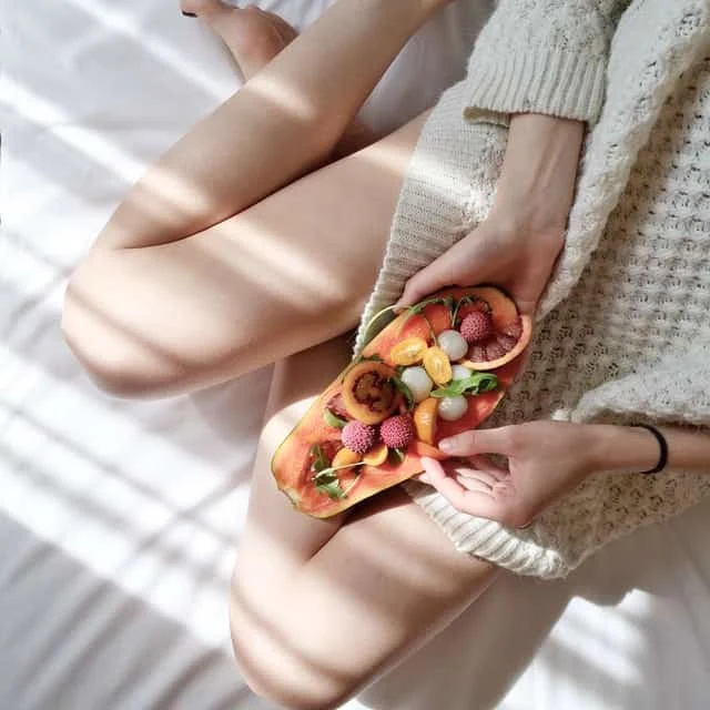A person in a cozy sweater sits on a bed with crossed legs, holding a colorful fruit-topped toast. Sunlight filters through blinds, casting soft shadows across the scene.