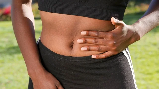 A person wearing a black crop top and pants stands outdoors, holding their side and stomach with one hand, possibly indicating discomfort or pain.