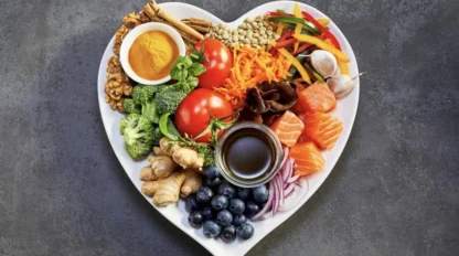 A heart-shaped plate filled with a variety of fresh foods including tomatoes, salmon, broccoli, blueberries, nuts, spices, ginger, garlic, and sliced vegetables.