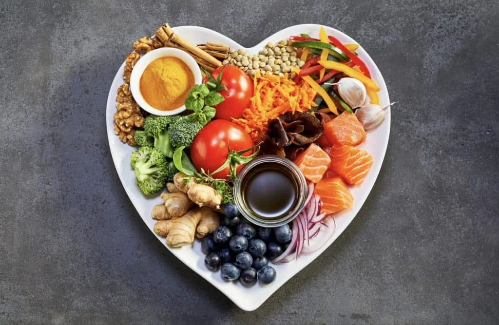 A heart-shaped plate filled with a variety of fresh foods including tomatoes, salmon, broccoli, blueberries, nuts, spices, ginger, garlic, and sliced vegetables.
