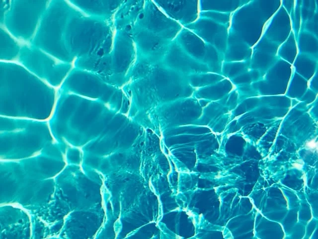 Sunlight reflects off the surface of clear blue water, creating bright, wavy patterns and ripple effects. The image captures the vibrant, shimmering appearance of water in a swimming pool or natural body of water.