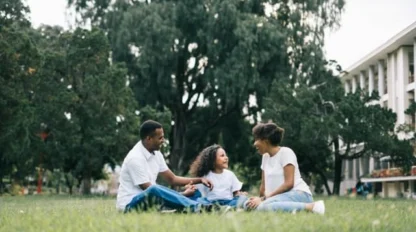Three people sit together on grass in a park, talking and smiling, with trees and a building in the background.