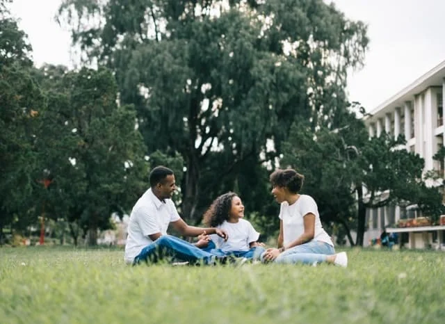 Three people sit together on grass in a park, talking and smiling, with trees and a building in the background.