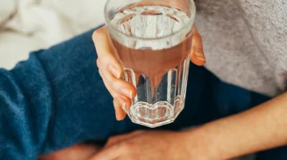 A person sitting on a bed, wearing blue jeans and a gray shirt, holds a clear glass of water.
