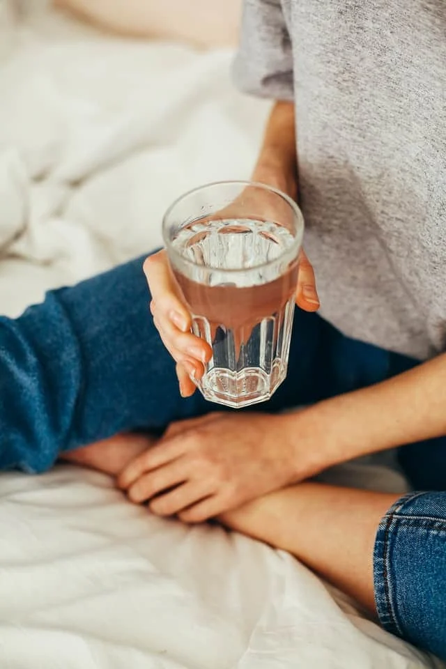 A person sitting on a bed, wearing blue jeans and a gray shirt, holds a clear glass of water.