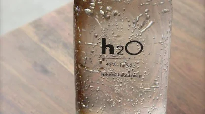 A clear glass bottle labeled "h2O" is filled with water and placed on a wooden surface next to a white sheet of paper.