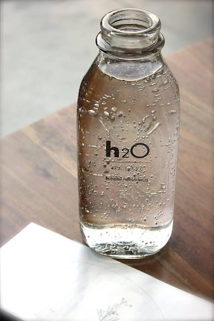 A clear glass bottle labeled "h2O" is filled with water and placed on a wooden surface next to a white sheet of paper.