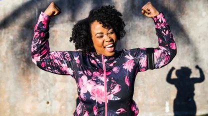 A woman in a floral athletic jacket smiles joyfully and flexes her arms in a strong pose outdoors, with her shadow showing the same pose on the wall behind her.