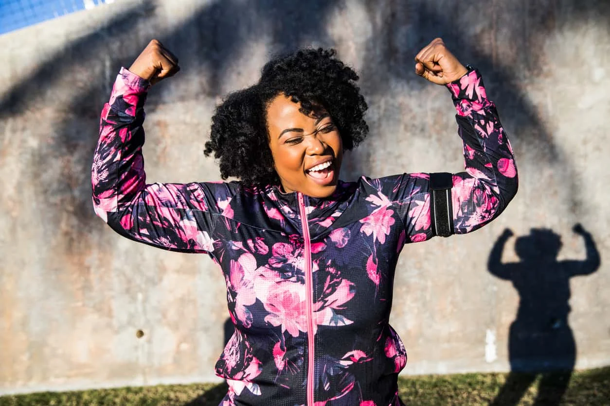 A woman in a floral athletic jacket smiles joyfully and flexes her arms in a strong pose outdoors, with her shadow showing the same pose on the wall behind her.