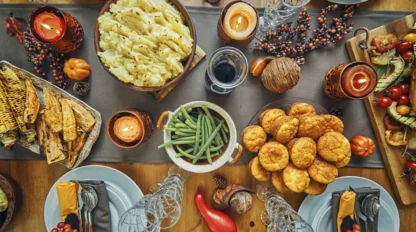 A festive table set for a meal with dishes including mashed potatoes, green beans, cornbread muffins, bread, vegetables, candles, and autumn decorations like mini pumpkins and berries. Four place settings are arranged around the table.