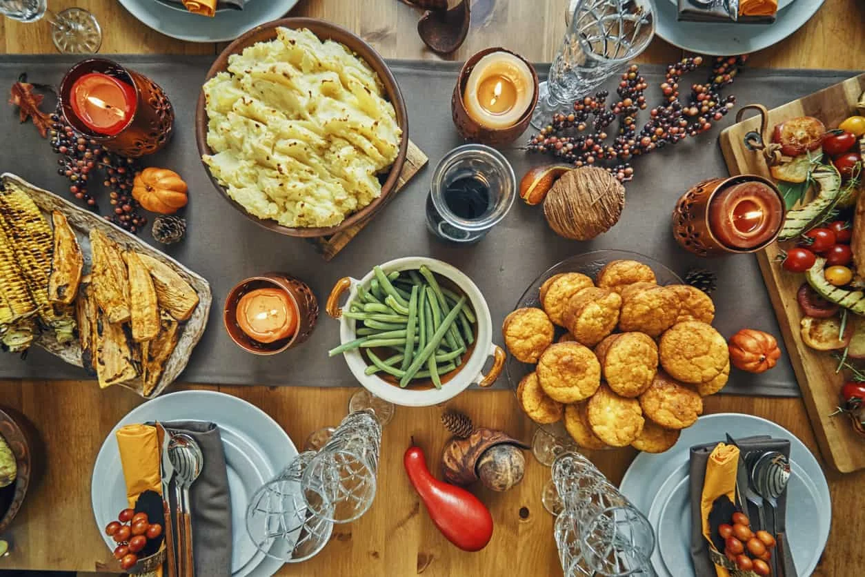 A festive table set for a meal with dishes including mashed potatoes, green beans, cornbread muffins, bread, vegetables, candles, and autumn decorations like mini pumpkins and berries. Four place settings are arranged around the table.