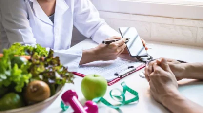 A nutritionist consults with a patient, pointing at a smartphone over a table with a salad, apple, pink dumbbells, and a measuring tape.