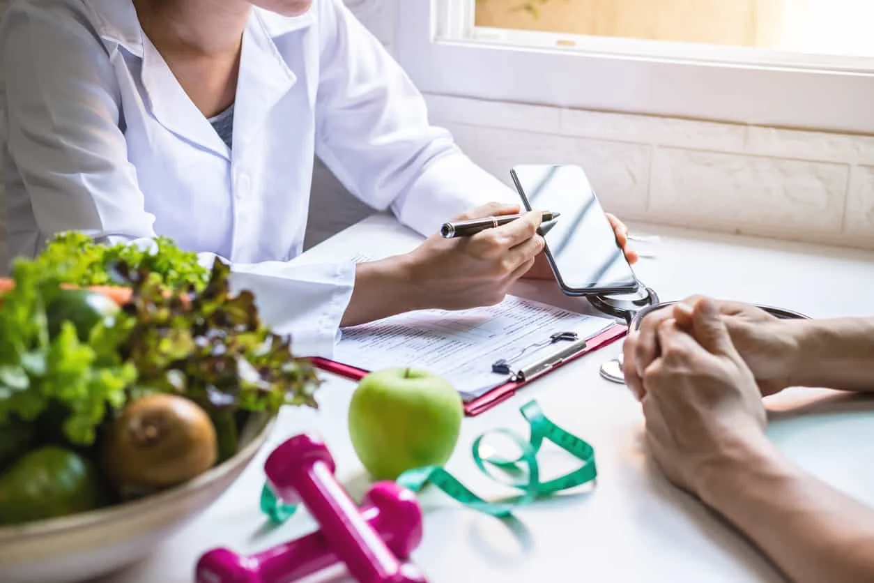 A nutritionist consults with a patient, pointing at a smartphone over a table with a salad, apple, pink dumbbells, and a measuring tape.