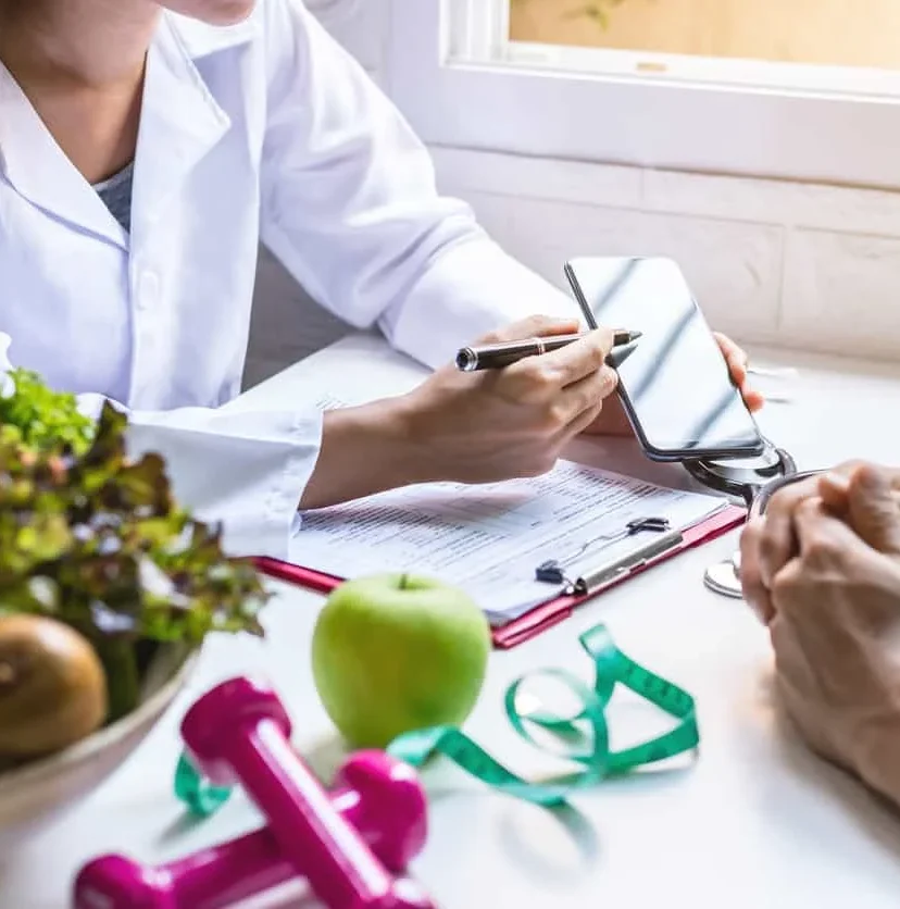 A nutritionist consults with a patient, pointing at a smartphone over a table with a salad, apple, pink dumbbells, and a measuring tape.