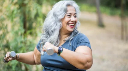 Smiling older woman with long gray hair exercises outdoors, holding small hand weights in both hands. She wears a teal shirt and a smartwatch, and is surrounded by greenery.