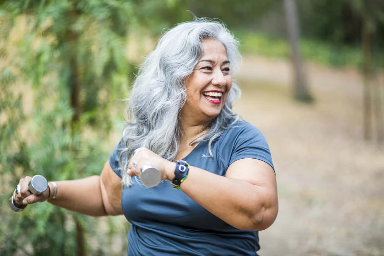 Smiling older woman with long gray hair exercises outdoors, holding small hand weights in both hands. She wears a teal shirt and a smartwatch, and is surrounded by greenery.