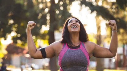 A woman in athletic wear smiles joyfully with her arms raised in victory while standing outdoors in a sunlit park, celebrating a fitness achievement.